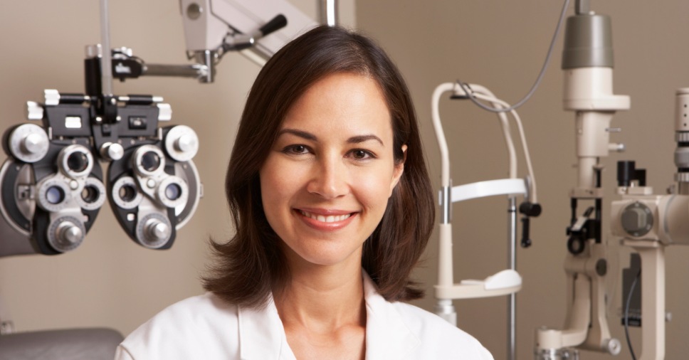 Médica sorridente em consultório equipado reforça a importância de ir ao oftalmologista.